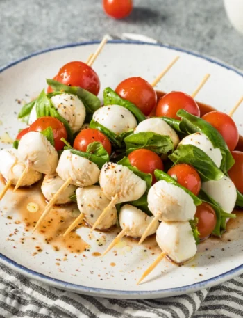 A white serving platter with colorful Caprese skewers neatly arranged. Each skewer features a bright red cherry tomato, a fresh green basil leaf, and a smooth white mozzarella ball, lightly drizzled with olive oil and balsamic glaze. The skewers are sprinkled with sea salt and black pepper, set on a light wooden table with natural lighting, highlighting the vibrant, fresh ingredients