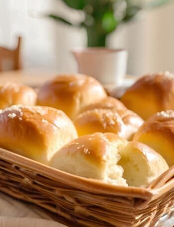 A basket of freshly baked, golden-brown dinner rolls topped with sesame seeds, arranged neatly and glistening with butter, in a cozy sunlit kitchen setting.